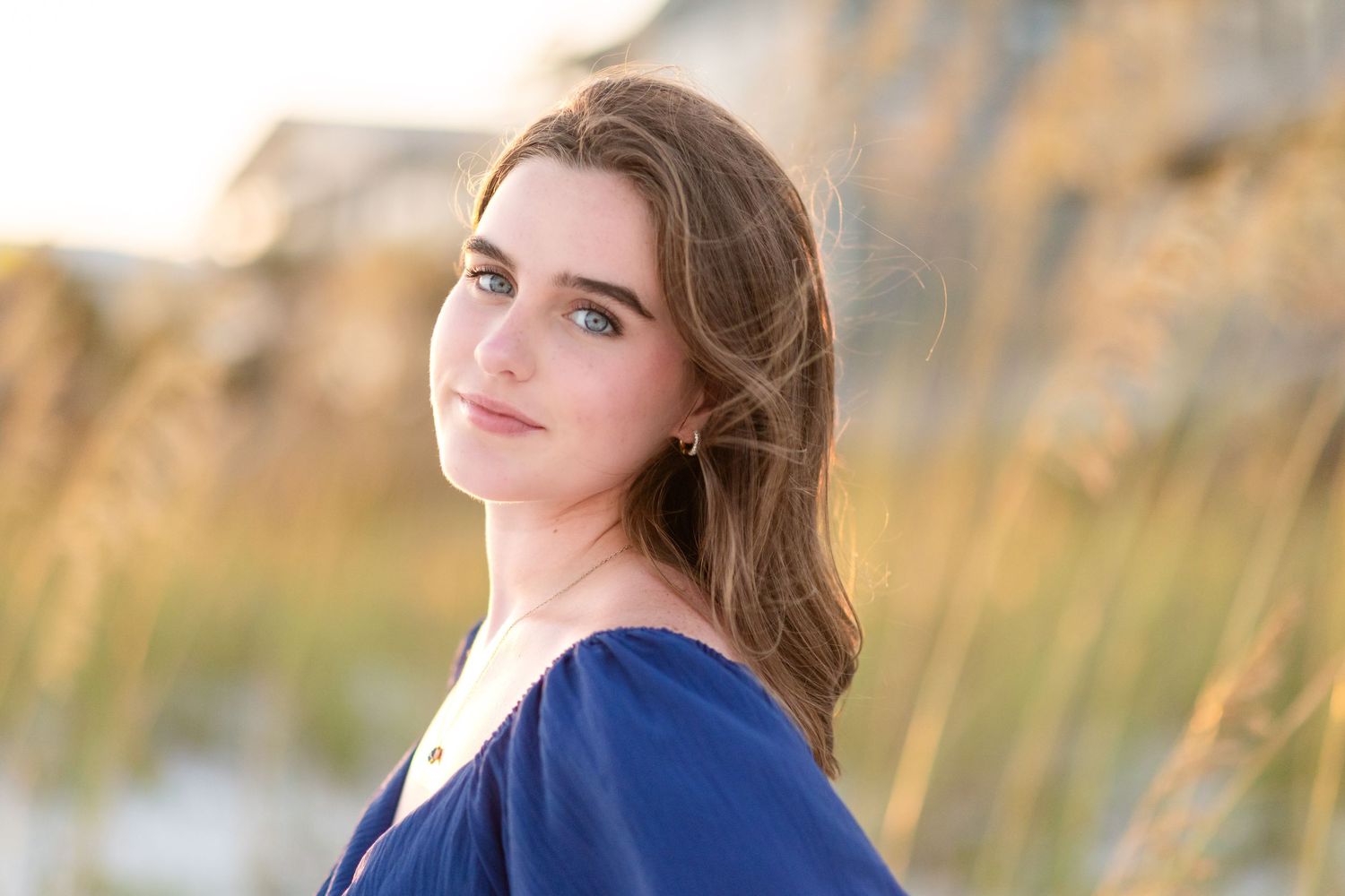 A young woman in a blue sweater poses outdoors with golden autumn grass and soft natural lighting in the background.