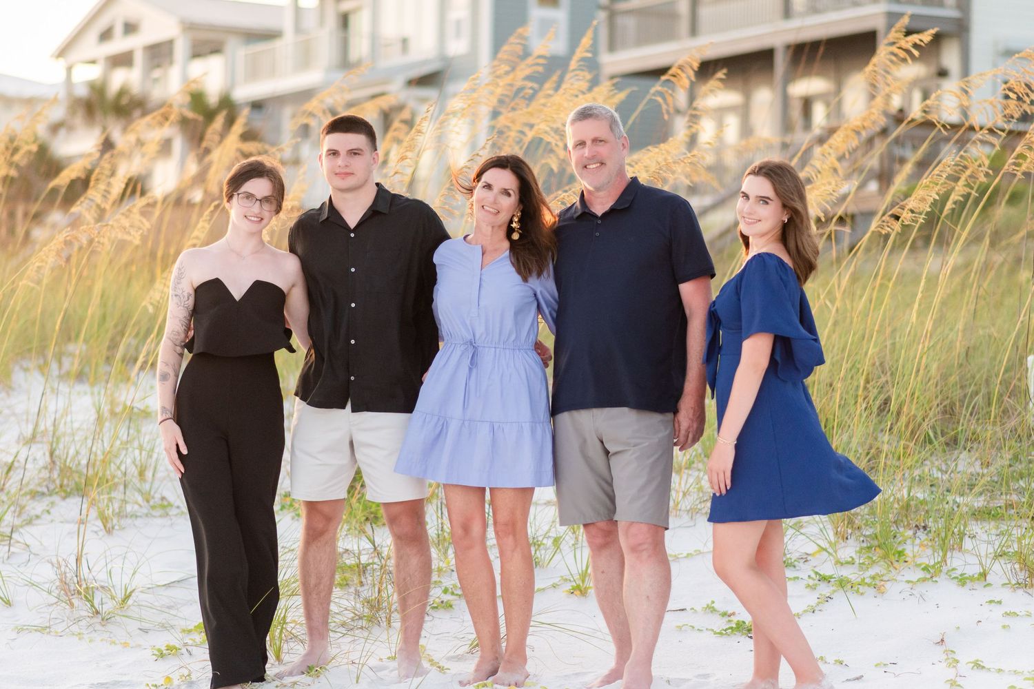A group of people in casual attire pose together on a sandy beach with dunes and beach grass in the background.