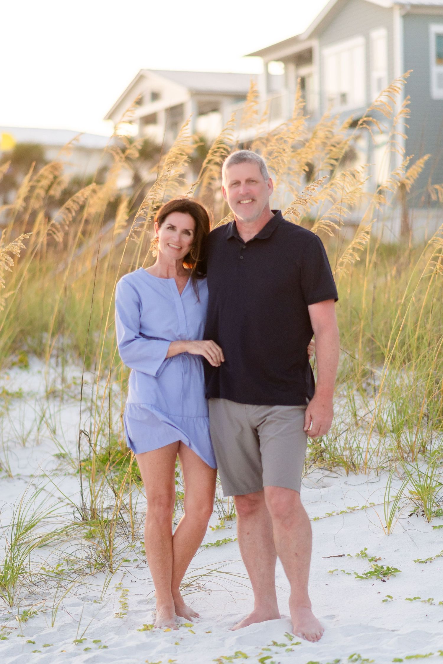 A couple stands together on a white sandy beach with sea oats and beach houses in the background at sunset.