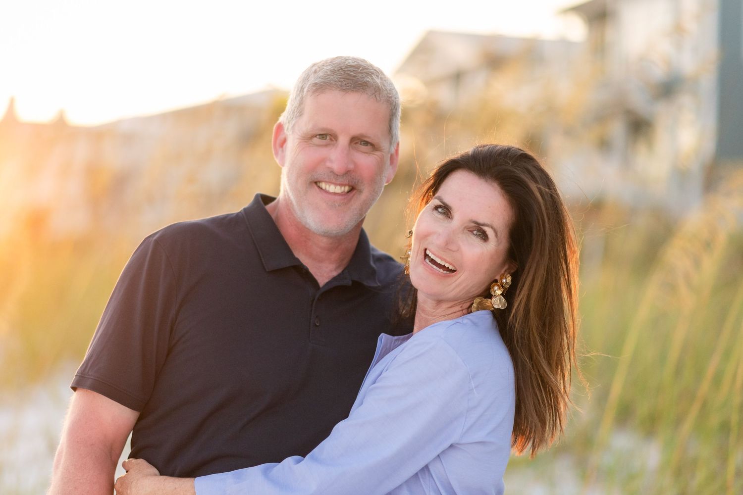 Couple embraces and smiles while posing outdoors during golden hour sunset in casual attire.