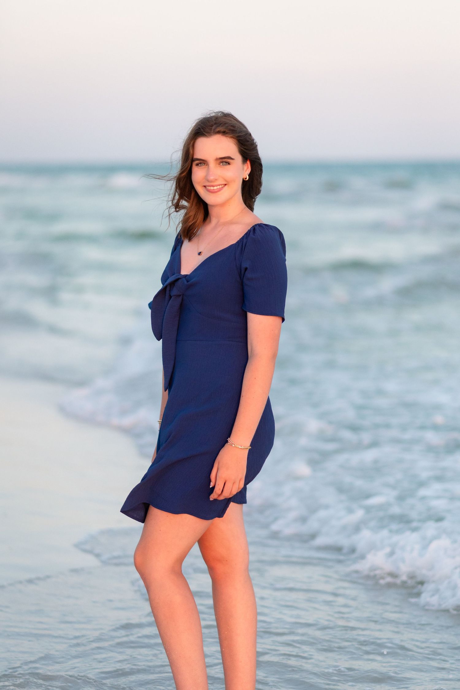 A person in a navy blue top poses playfully at the beach during sunset with ocean waves in the background.