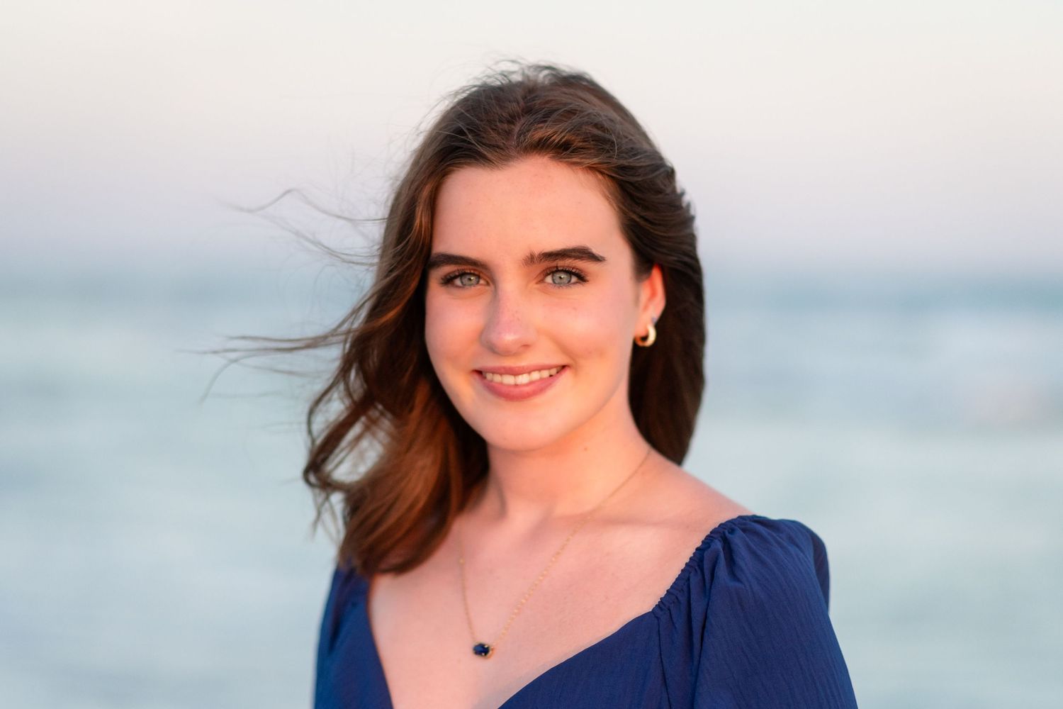 A person in a navy blue top poses outdoors with the ocean and cloudy sky visible in the background.