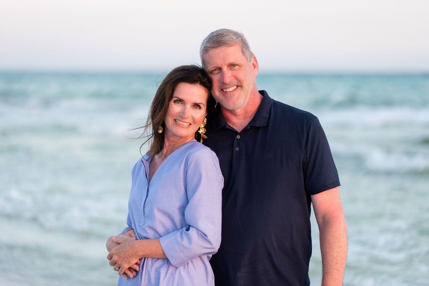 Couple poses together on a beach with ocean waves and blue sky in the background.