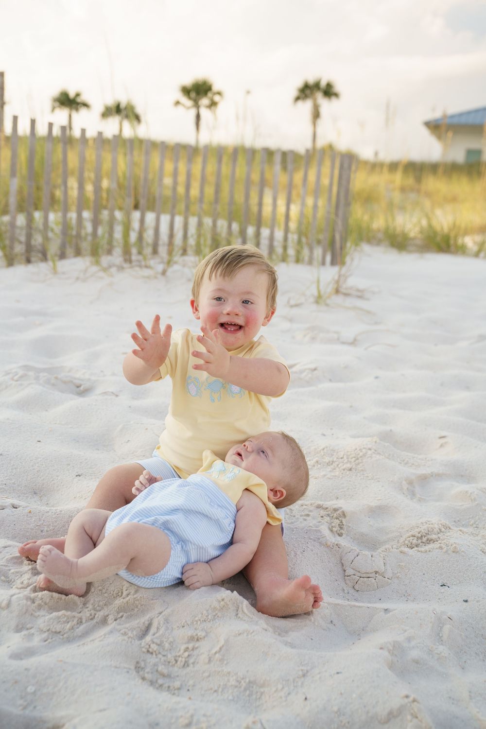 Two small children play together on white sandy beach with palm trees and sand fences in background.