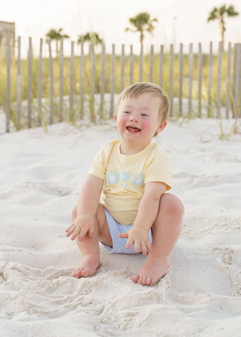 A happy toddler in yellow and blue plays in the soft white sand at the beach with palm trees in the background.