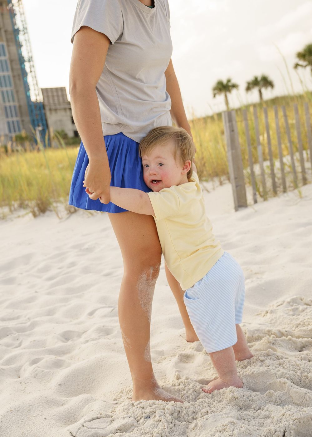 A toddler takes their first steps in soft beach sand while being supported by an adult.