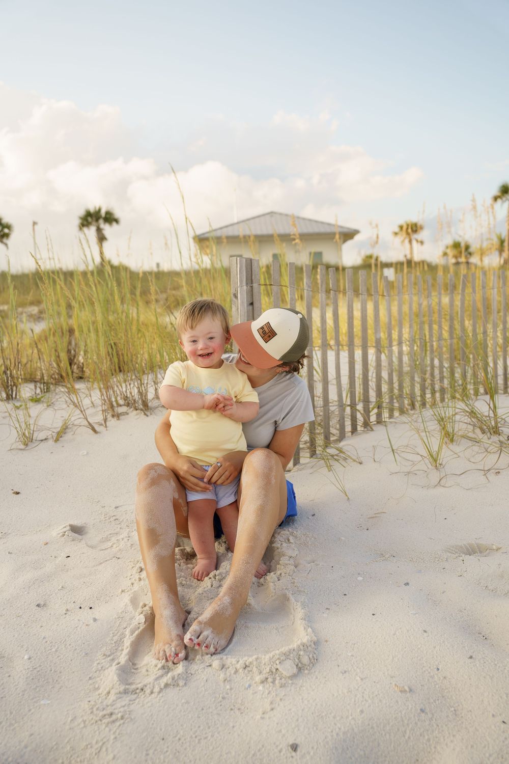 A heartwarming beach scene with two people sitting in the sand near palm trees and a coastal fence.