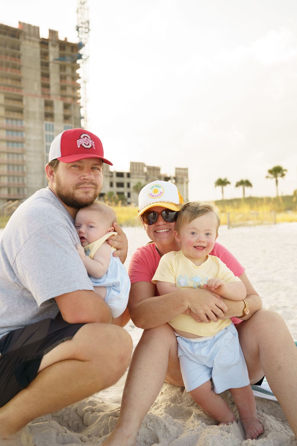 Family sitting together on beach sand at sunset with high-rise buildings visible in background.