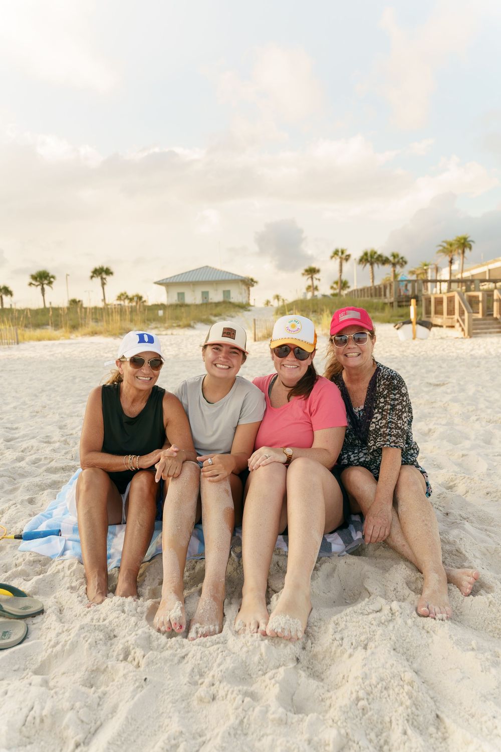 Friends relaxing on beach sand wearing casual summer attire and hats while posing for photo.