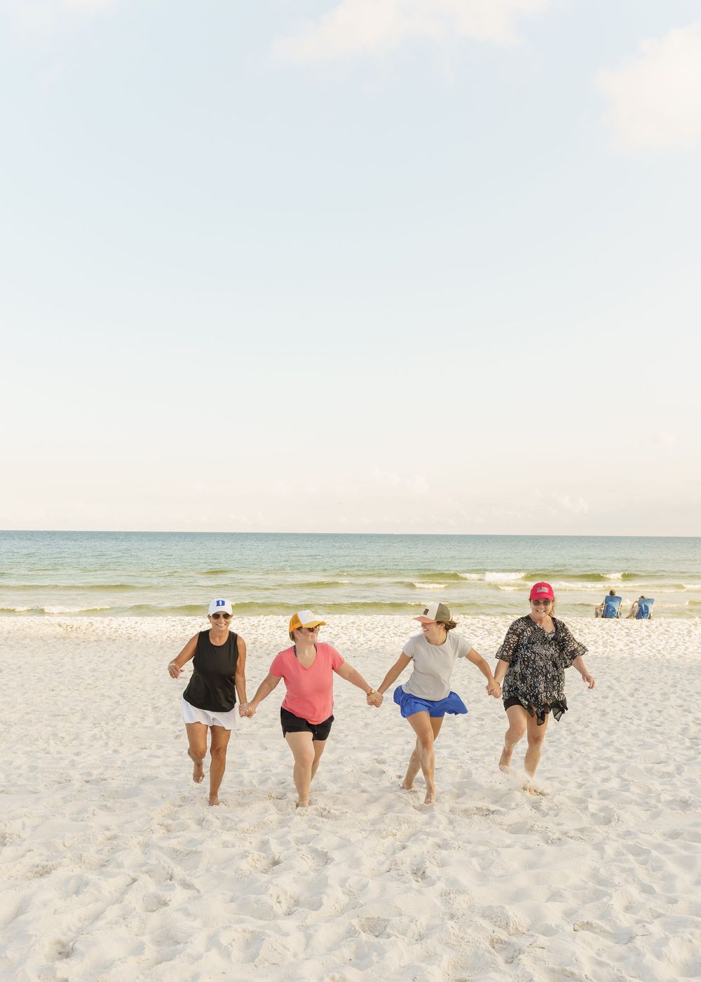 Four people holding hands while running joyfully along the shoreline on a sunny beach day.