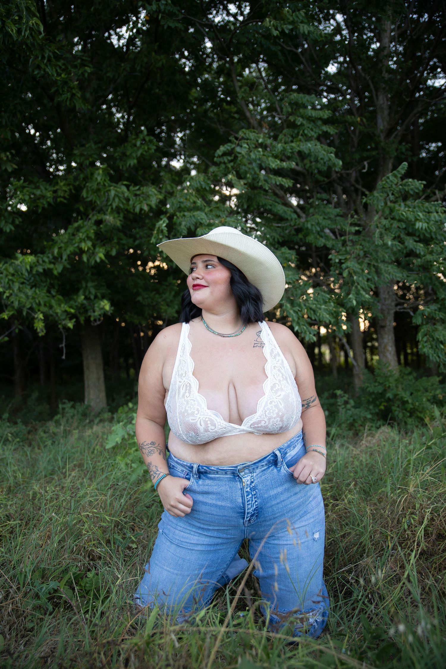 Someone in a white cowboy hat and lace top poses confidently in a field with trees in the background.