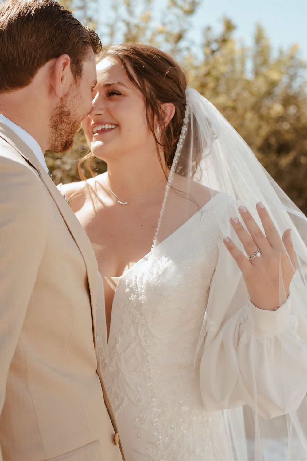 Romantic wedding couple embracing in soft sunlight with the bride wearing a white lace gown and flowing veil.