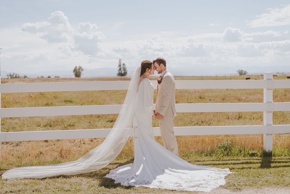 Wedding couple poses along white fence in open field with flowing bridal veil and natural landscape.