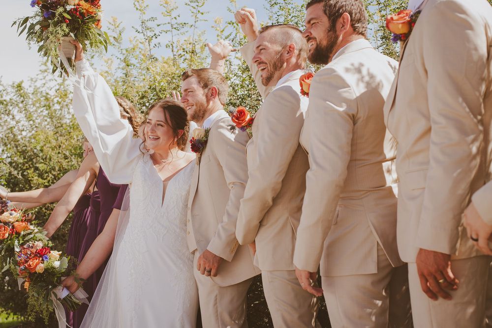 Wedding party in beige suits and white dresses lined up outdoors with colorful floral bouquets.
