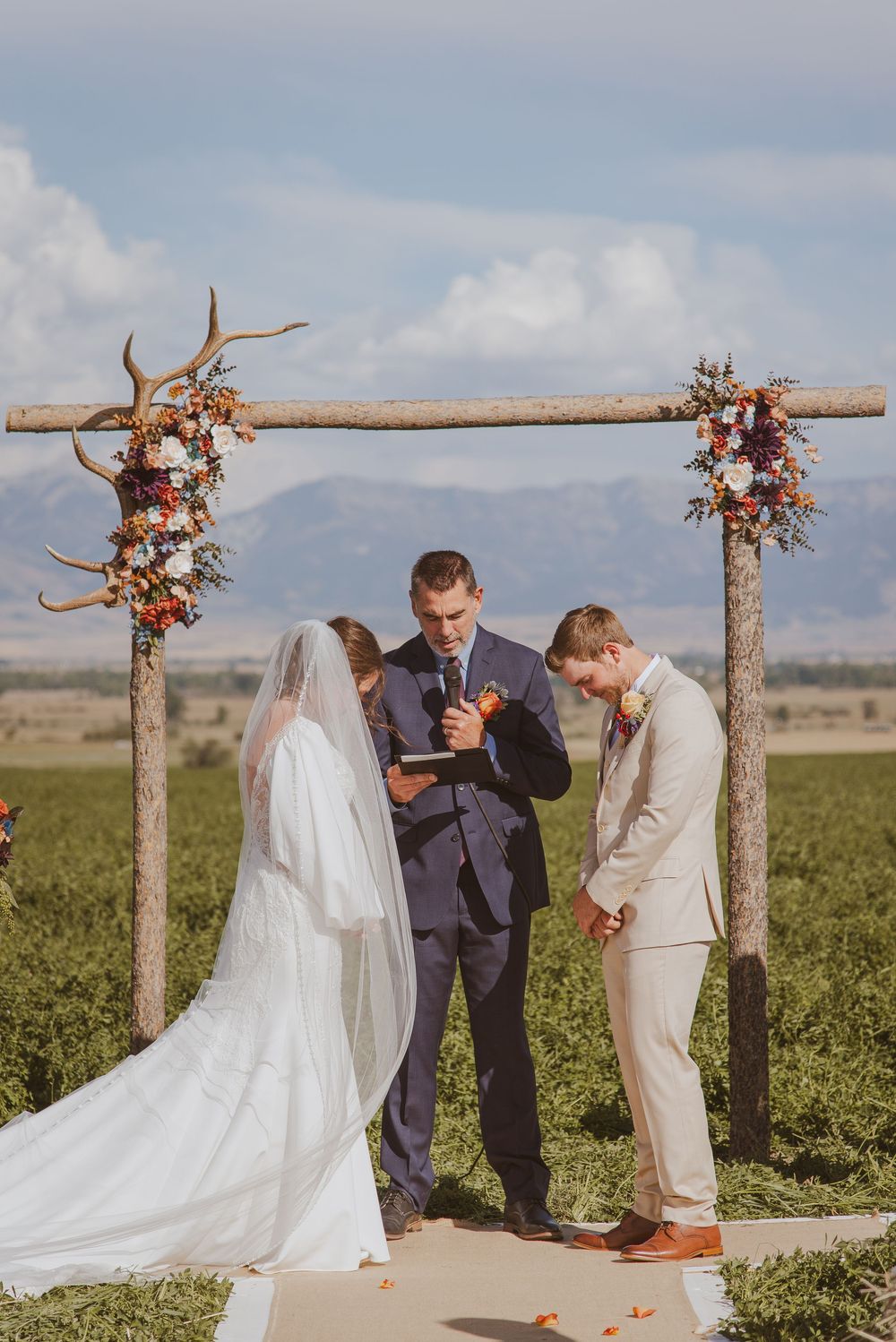 Wedding ceremony under rustic wooden arch decorated with flowers in outdoor field setting.