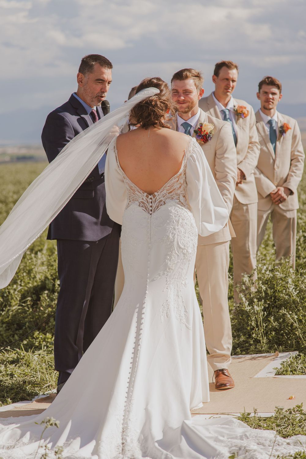 Wedding ceremony outdoors with bridal party in beige suits and flowing white dress against a natural landscape.