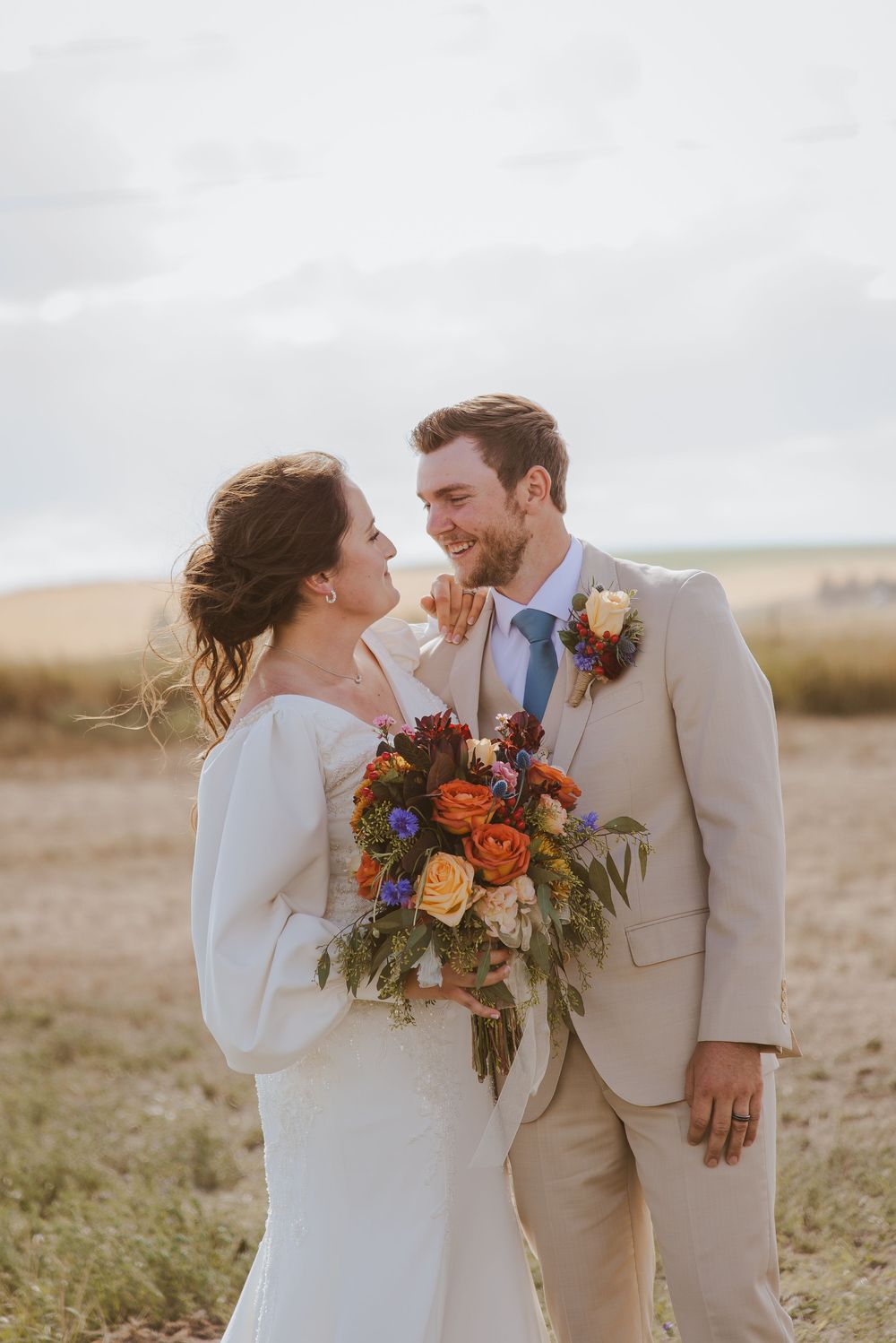 A joyful couple shares an intimate moment in a field during their wedding, with the bride holding a vibrant autumn bouquet.