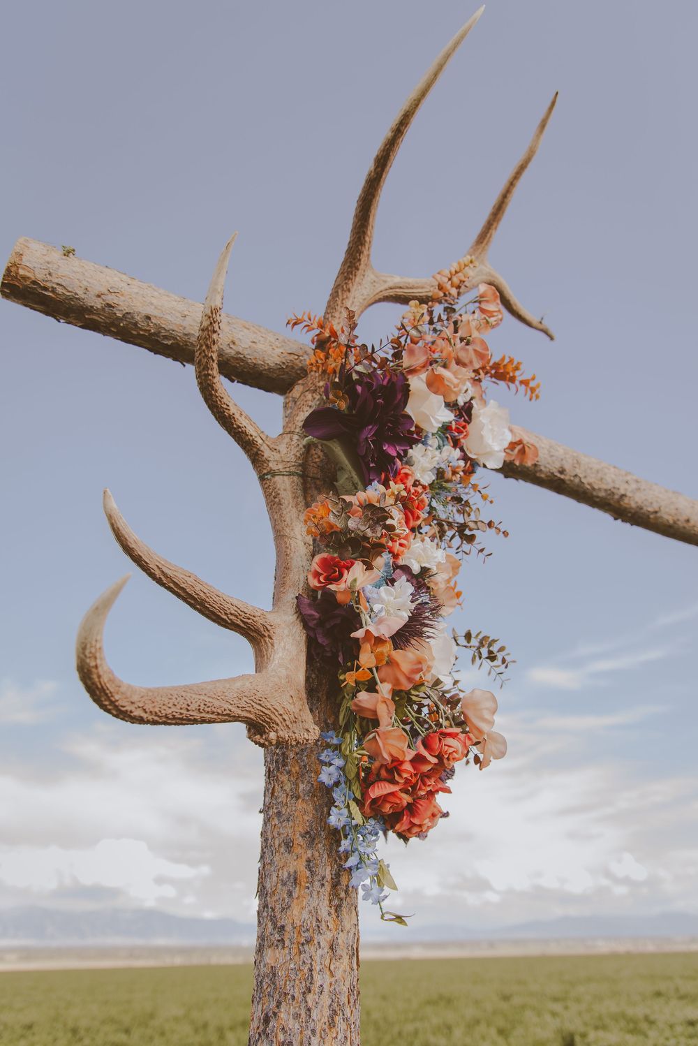 Rustic wooden arch decorated with orange and pink florals against a blue sky in an open field.