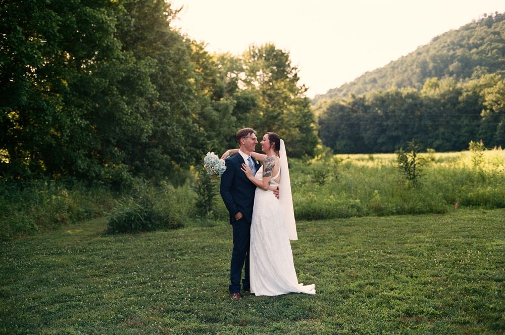 Bride and groom share a romantic kiss in a lush green meadow with rolling hills and trees at golden hour.