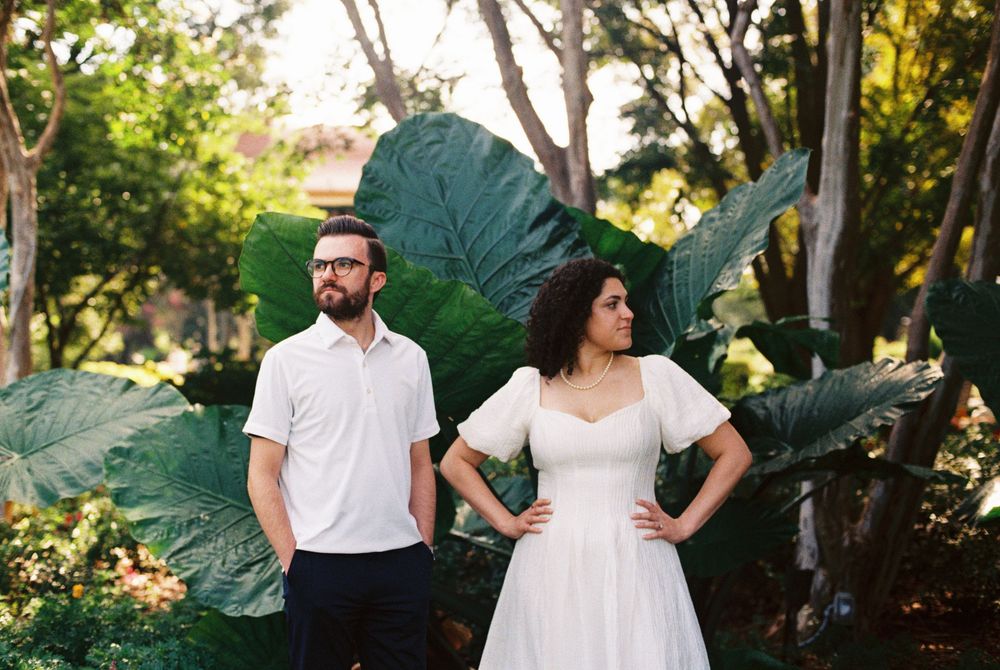 A couple walks hand in hand through a lush garden setting with large decorative plants during golden hour lighting.