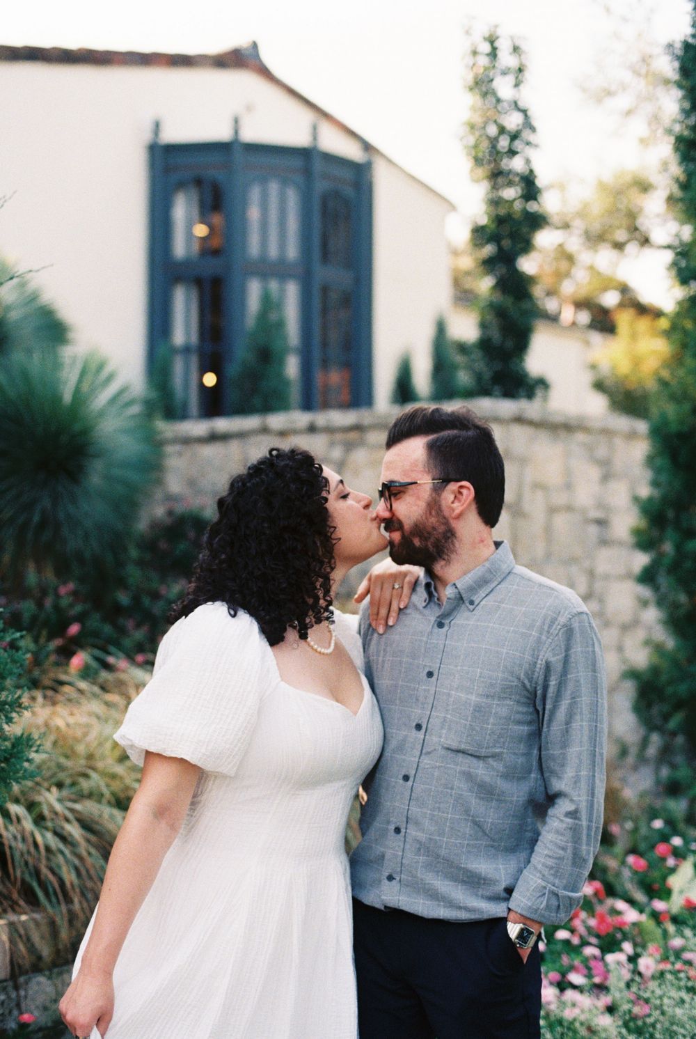 A couple shares an intimate moment in front of a white building with blue doors and garden landscaping.