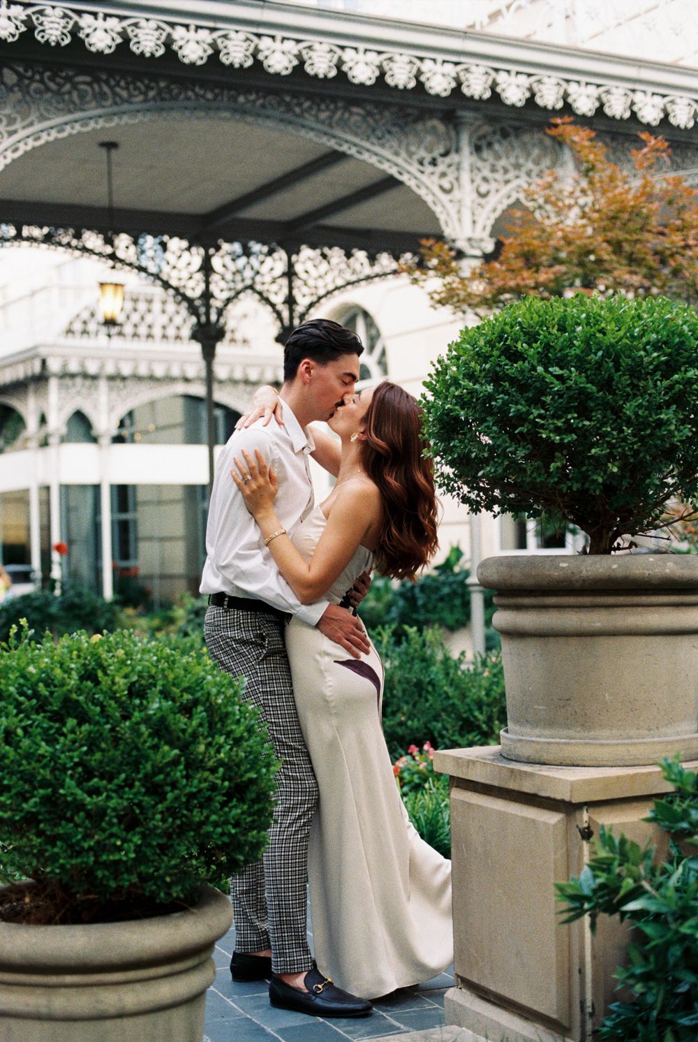 A romantic couple shares intimate moments on an elegant veranda with ornate architectural details and lush greenery.