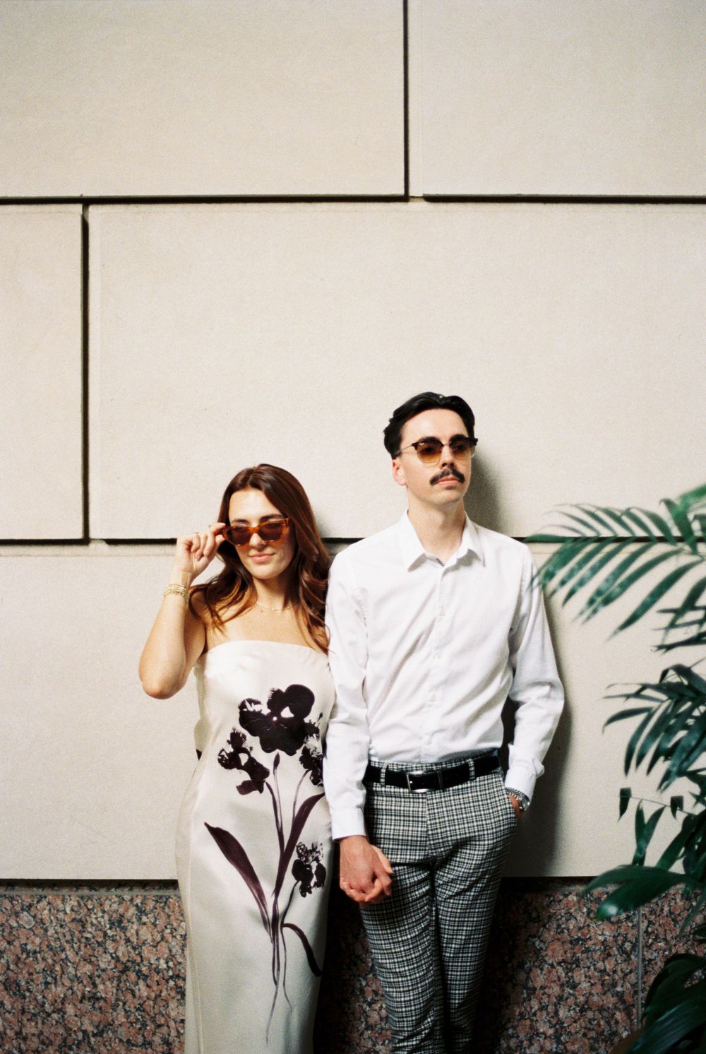 A couple in stylish attire poses against a white wall with plaid pants and a white dress near tropical plants.