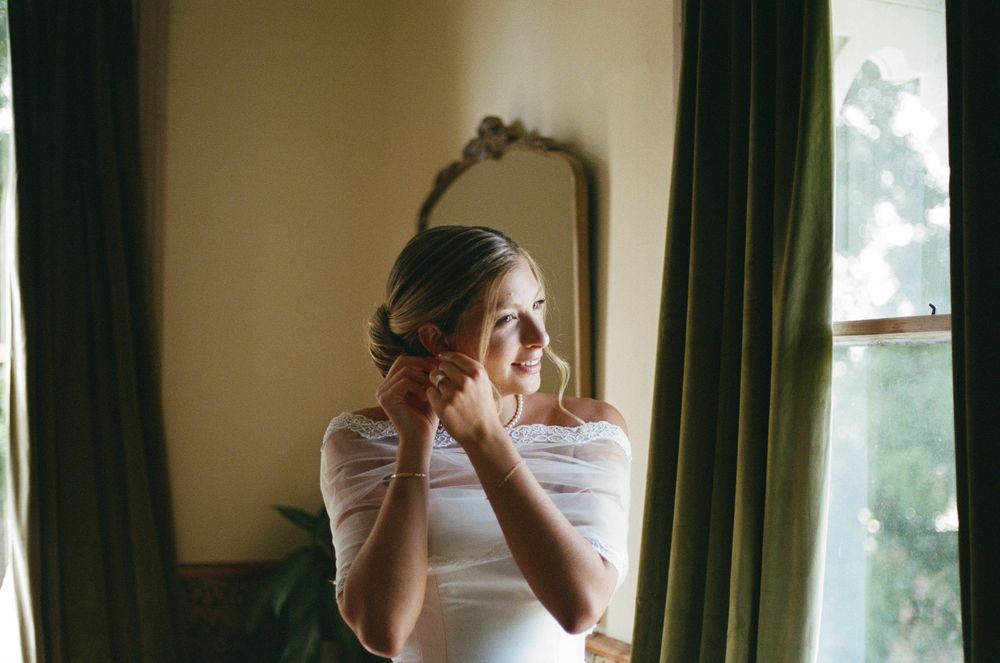 Bride in white dress adjusting earring by window with mirror, soft natural light, elegant wedding preparation moment.