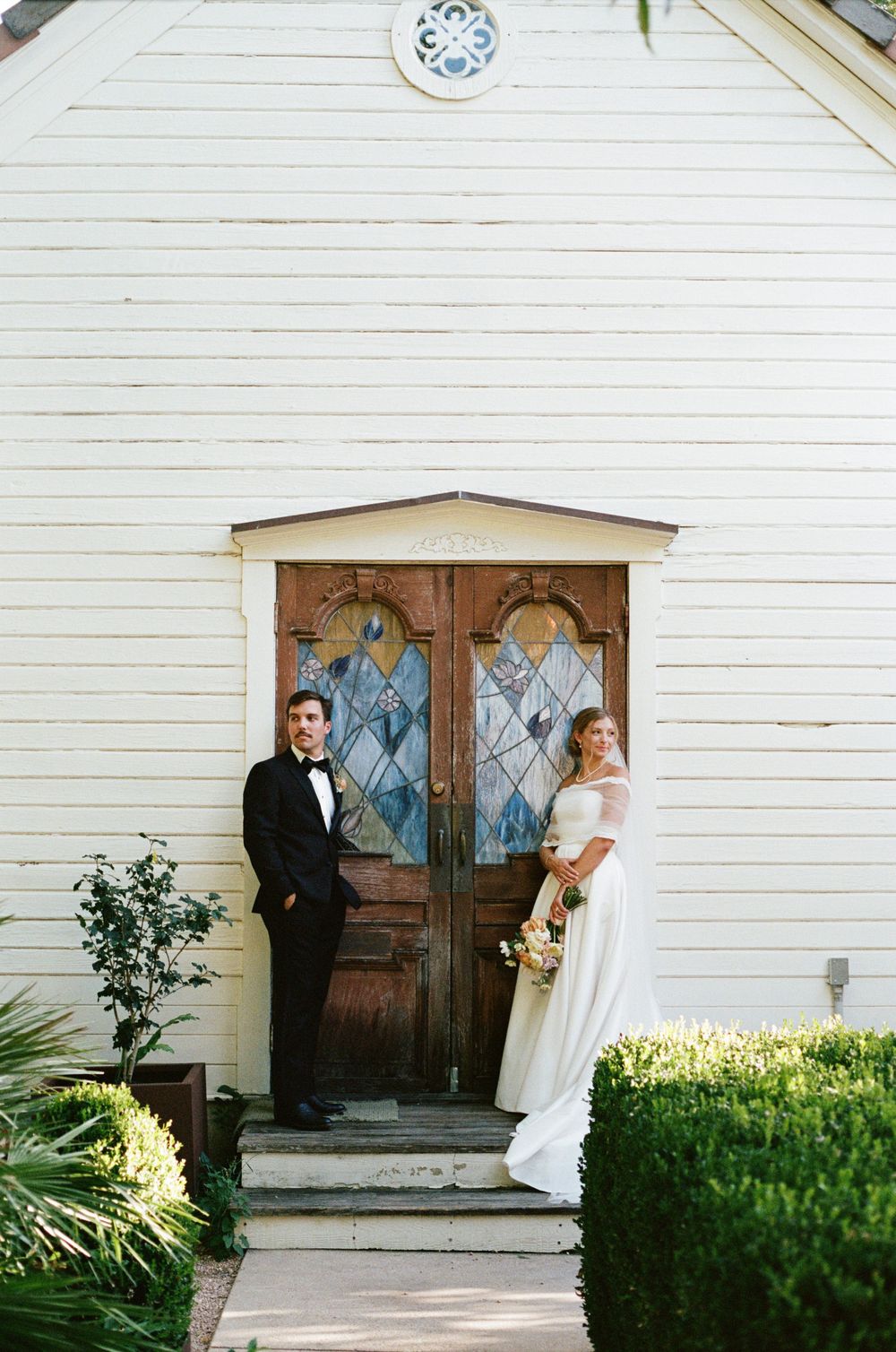Bride and groom pose by ornate wooden chapel doors with stained glass on their wedding day.