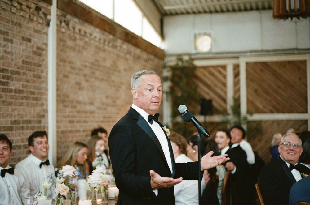 Man in tuxedo giving wedding speech at microphone in rustic brick venue with guests seated at decorated tables.