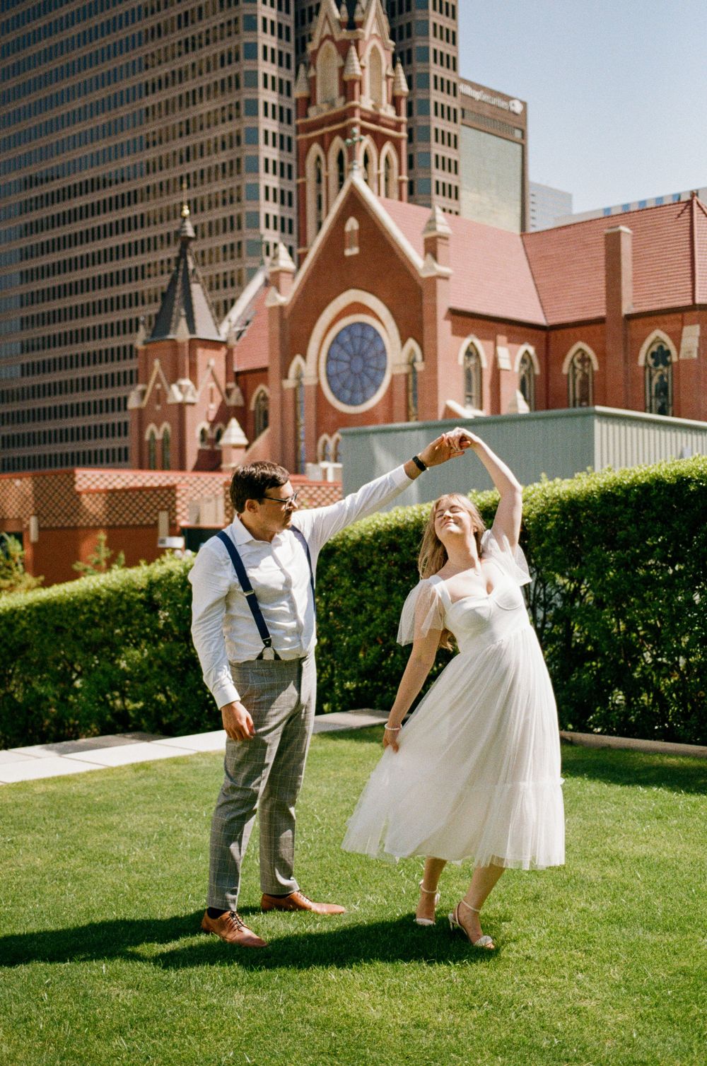 A couple dances on a green lawn with a red brick church and city skyscrapers in the background on their wedding day.