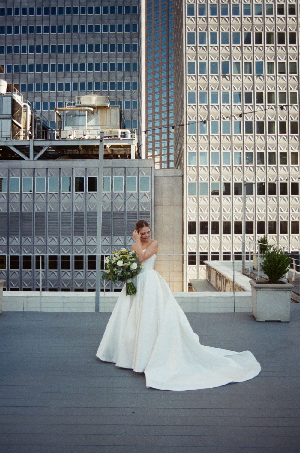 A bride in a flowing white gown poses dramatically on a modern rooftop terrace with city buildings in the background.