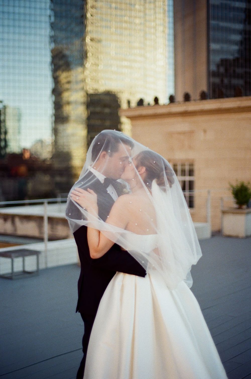 A romantic wedding couple embraces under a flowing white veil against an urban backdrop with tall buildings.