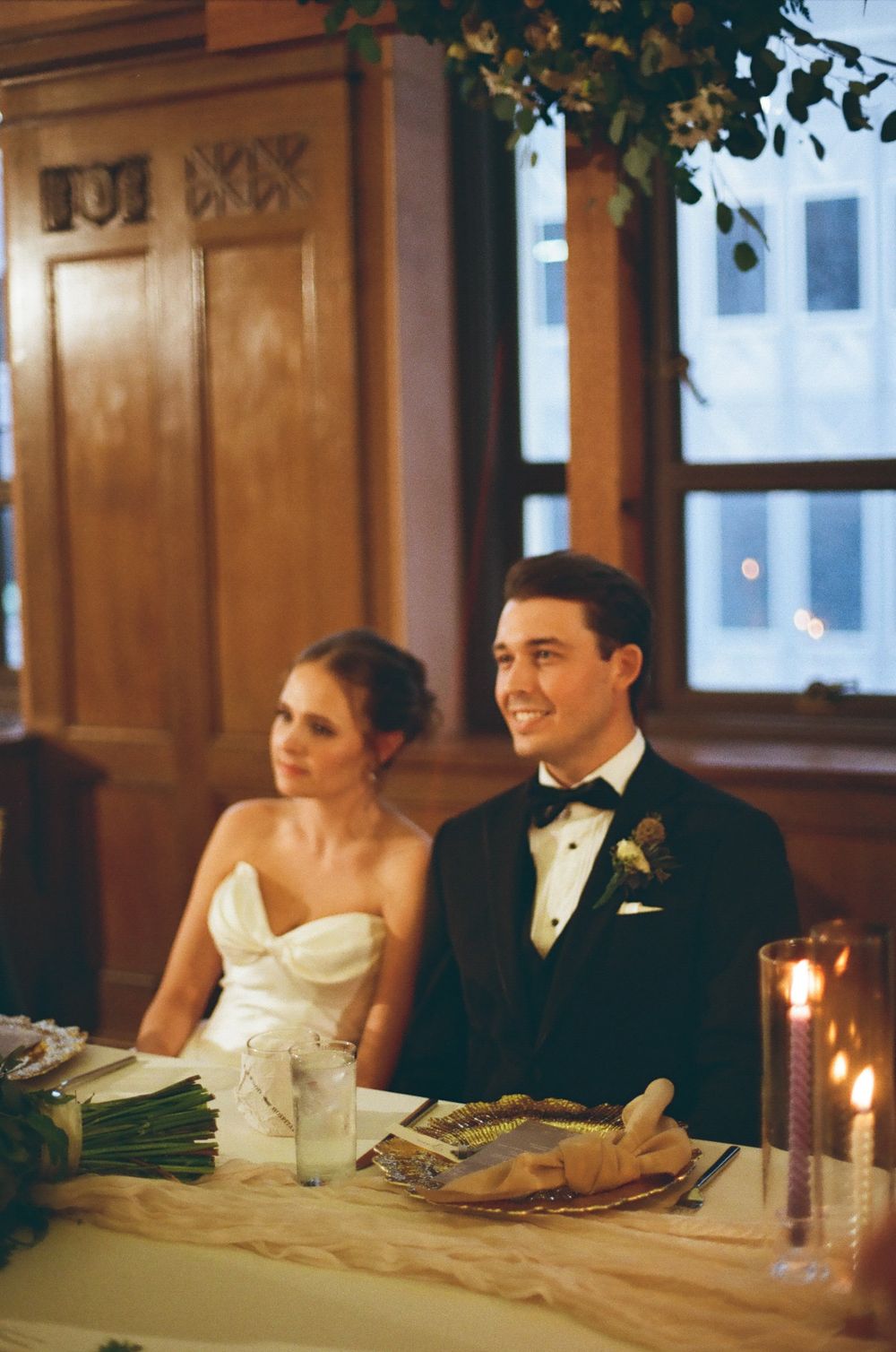 A couple in formal wedding attire sits together at a candlelit reception table, sharing an intimate moment.