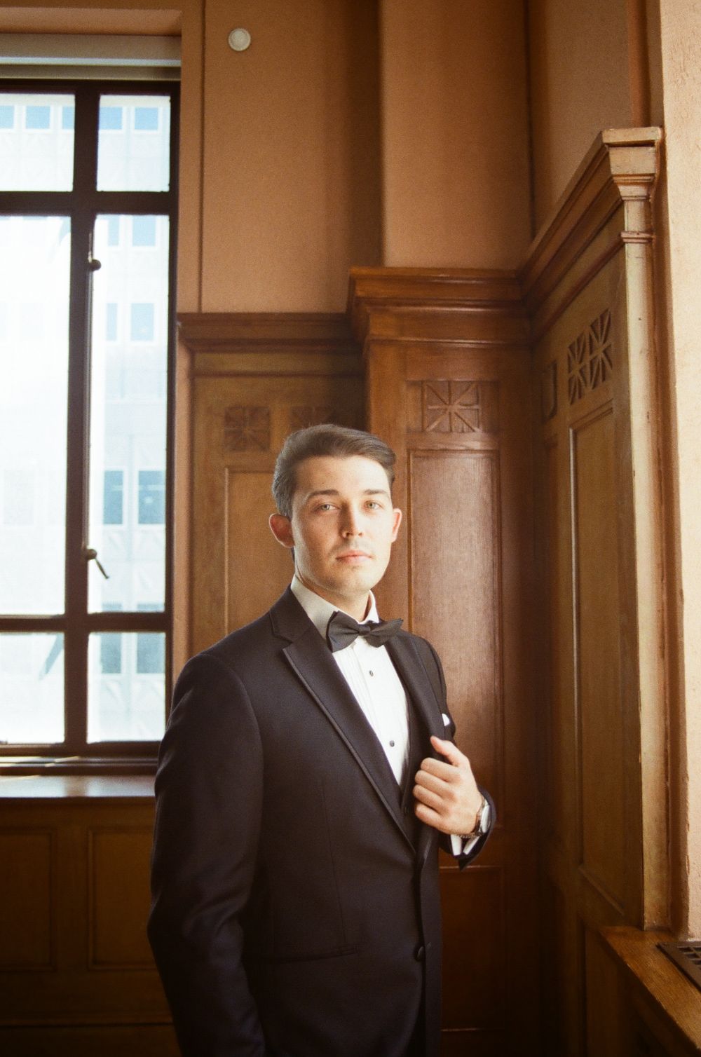 Professional portrait of a person in a dark suit standing in an elegant wood-paneled hallway with tall windows.