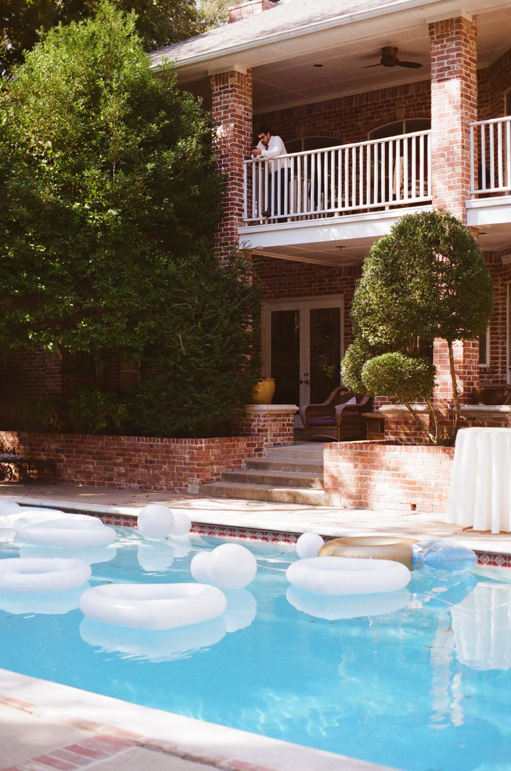 Swimming pool with inflatable floats surrounded by brick building and lush greenery on a sunny day.