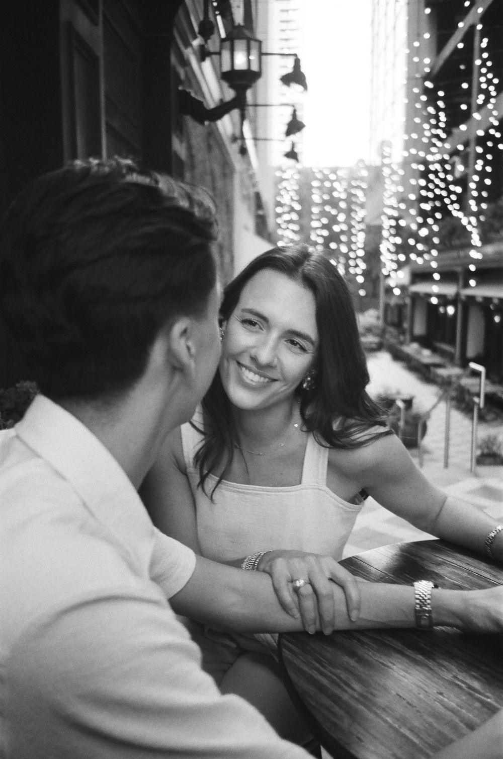 A romantic couple shares a tender moment at a restaurant table with twinkling lights in the background.