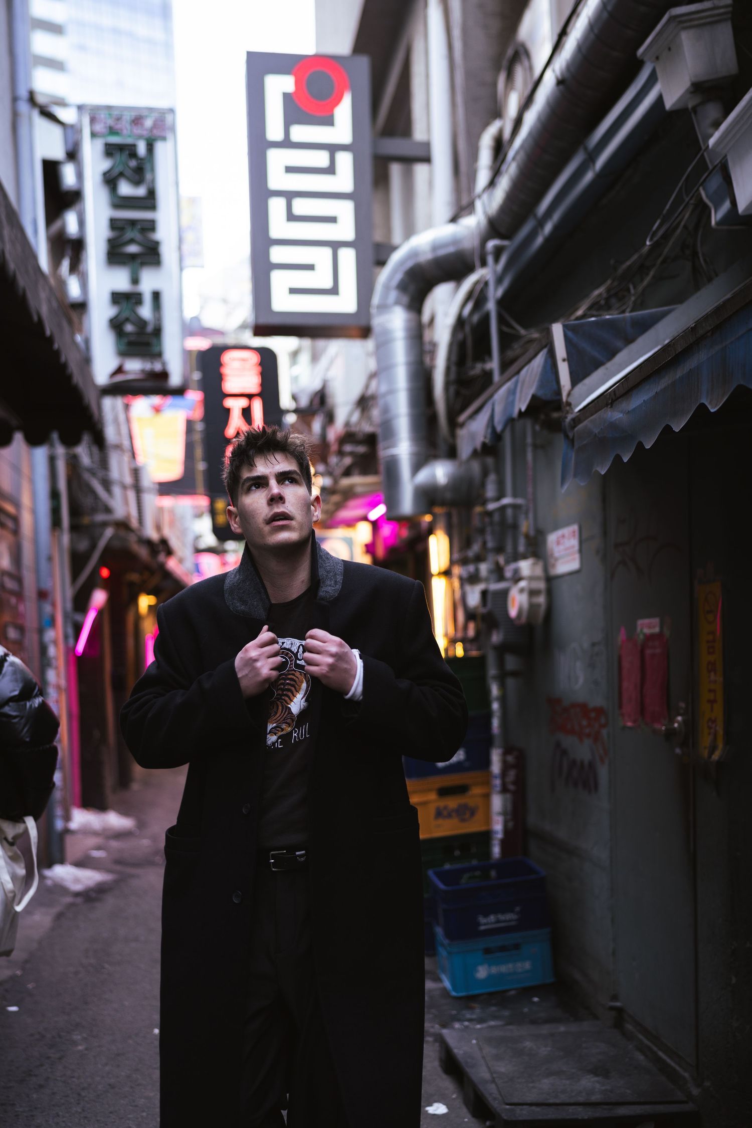 Person in dark clothing stands in dimly lit urban alley with neon lights glowing in the background.