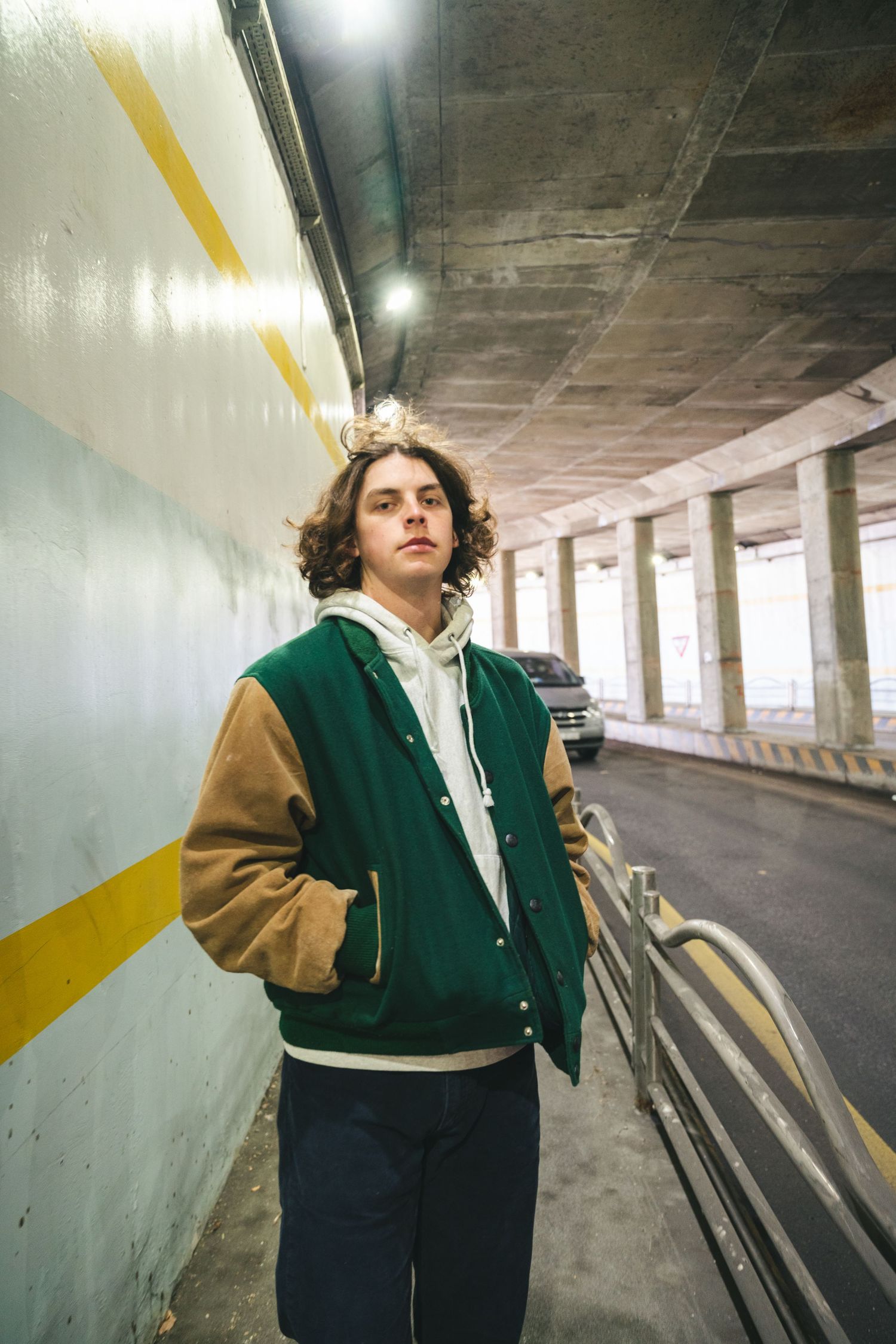 A person wearing a two-tone varsity jacket poses against a concrete wall in an empty parking garage.