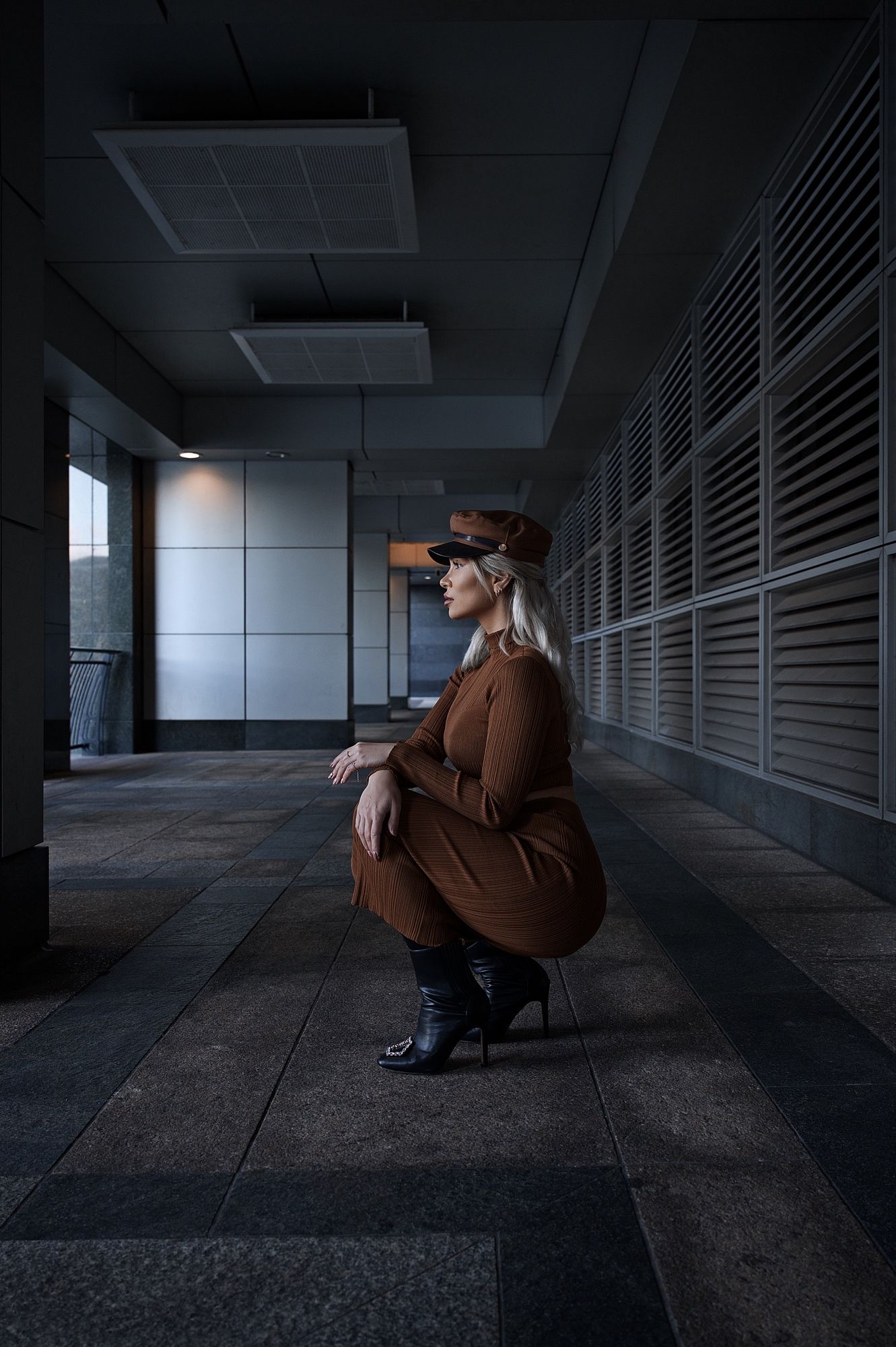 A person in a brown coat sits crouched in a dimly lit industrial hallway with concrete floors and metal walls.