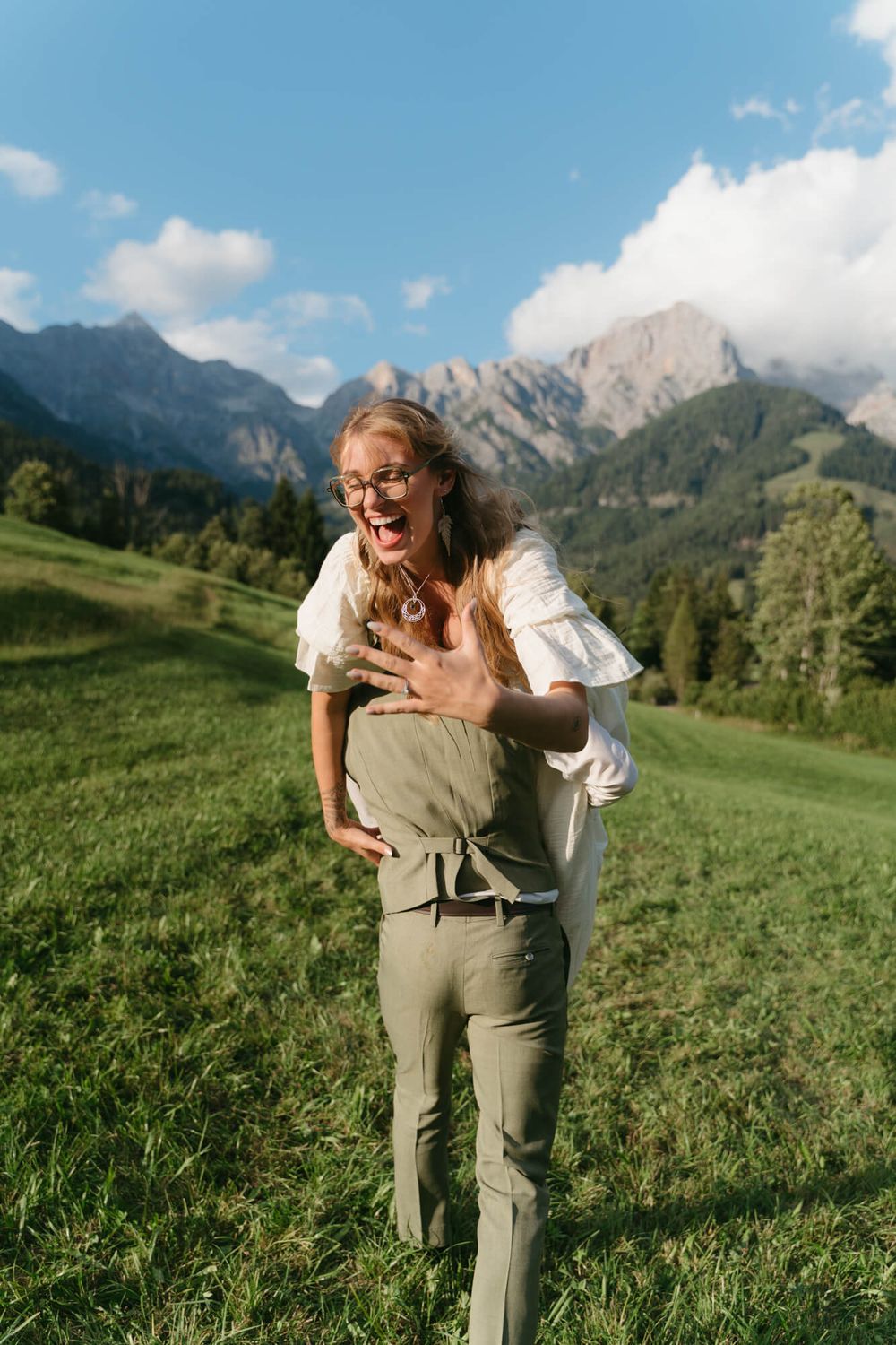 A playful couple shares a joyful moment in a scenic mountain meadow with dramatic alpine peaks in the background.