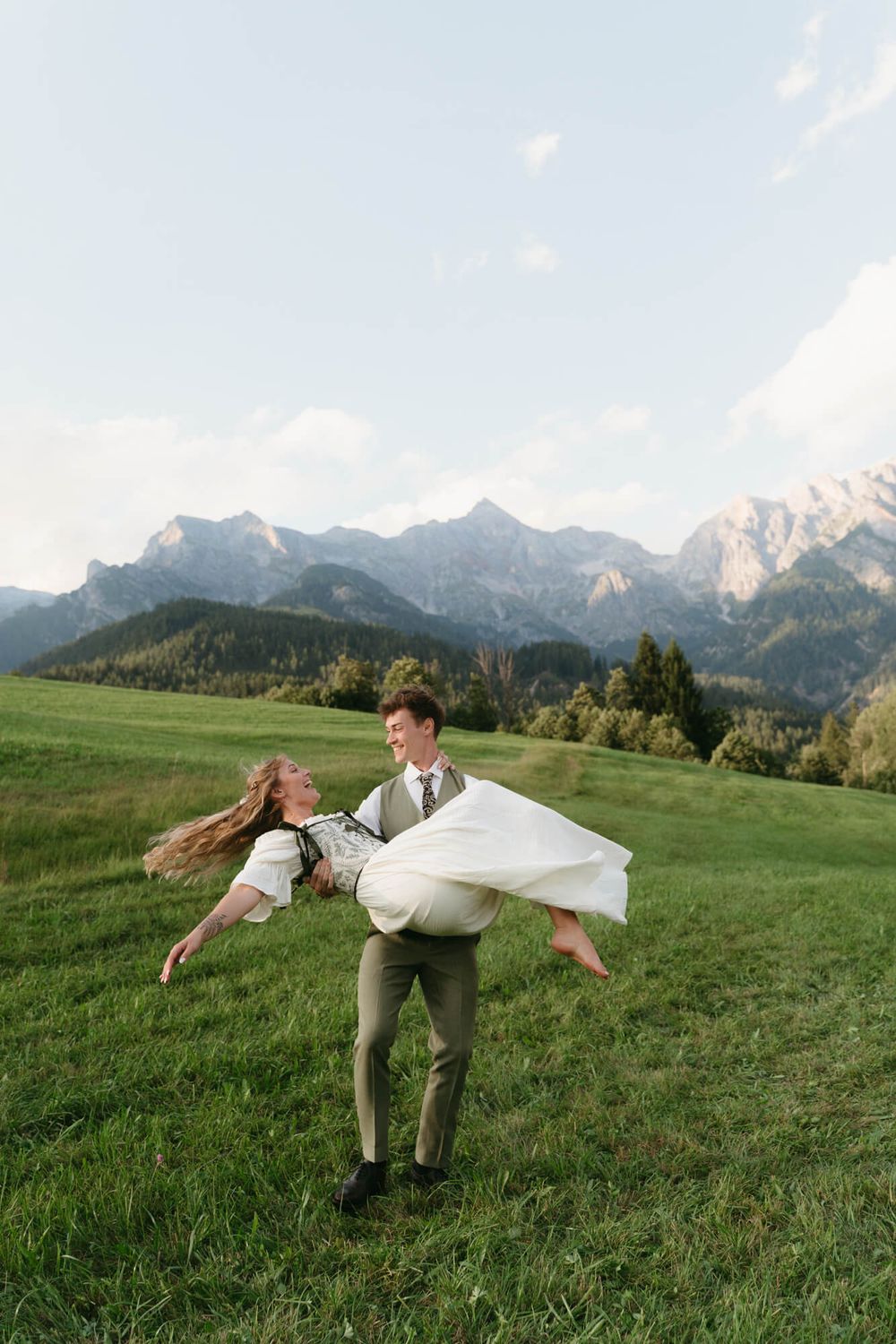 A couple playfully spins and dances in a green meadow with majestic mountain peaks in the background.