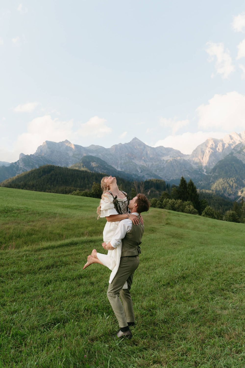 A couple embraces in a scenic mountain meadow with snow-capped Alpine peaks in the background.