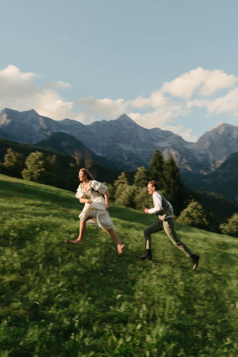 Joyful couple runs and leaps through a mountain meadow with dramatic alpine peaks in the distance.