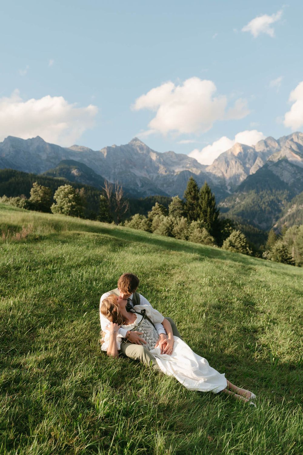Newlyweds relax together on a verdant alpine meadow with snow-capped mountains in the distance.