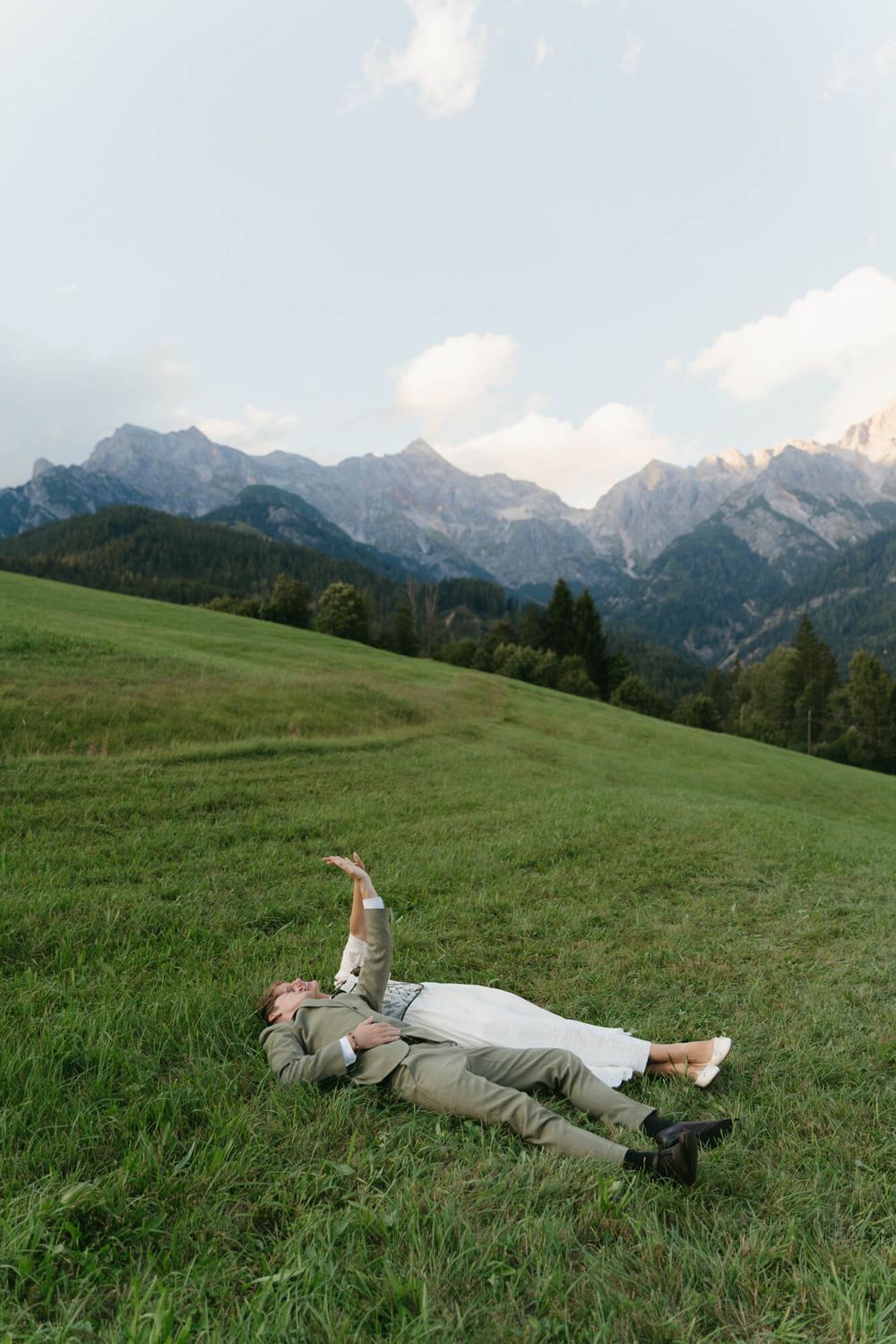 A person lies peacefully on a lush green hillside meadow with dramatic mountain peaks rising in the background.