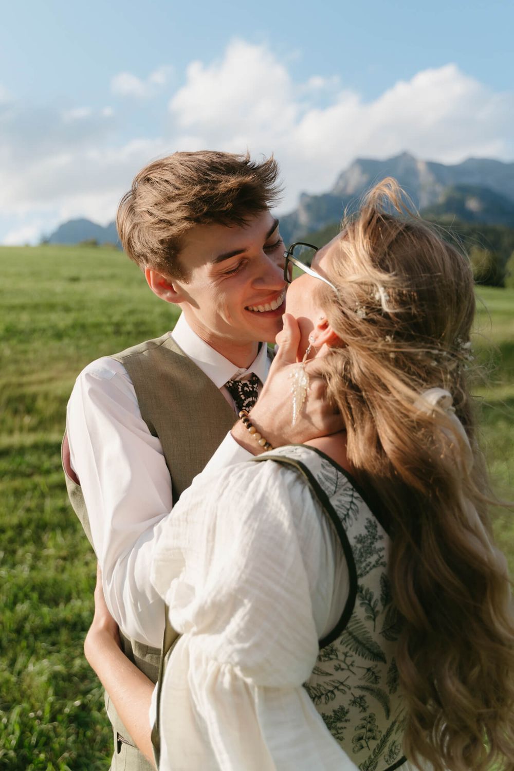A couple shares joyful moments together in a scenic mountain meadow with green grass and blue skies.