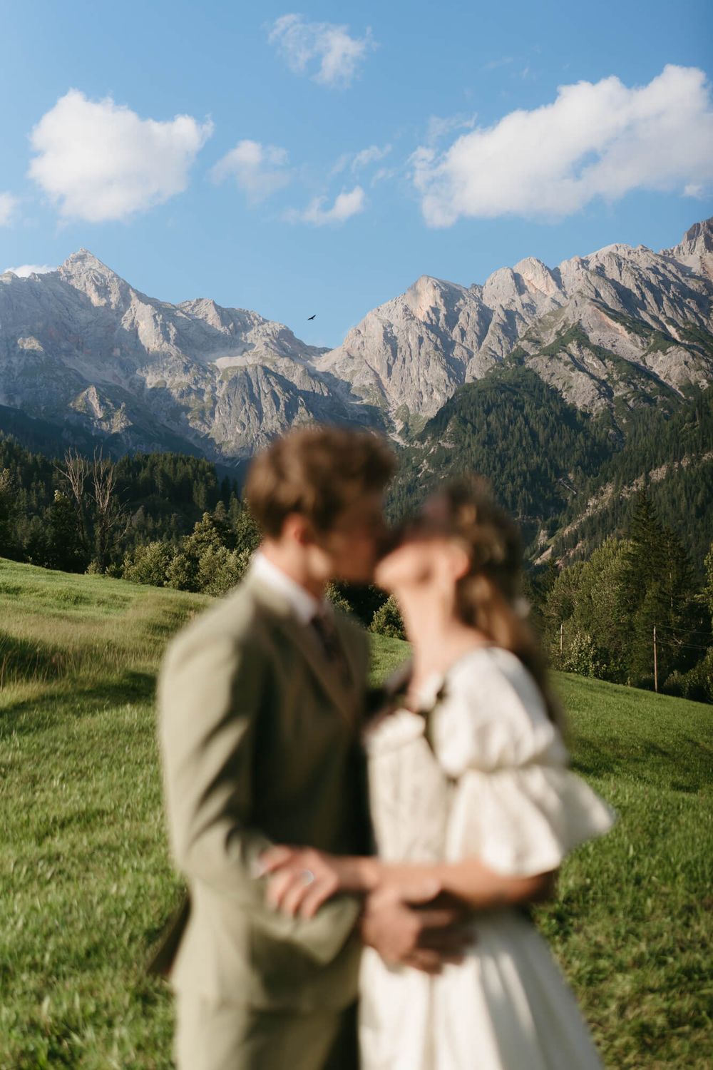 A couple shares an intimate moment in a scenic mountain meadow with dramatic alpine peaks rising in the background.