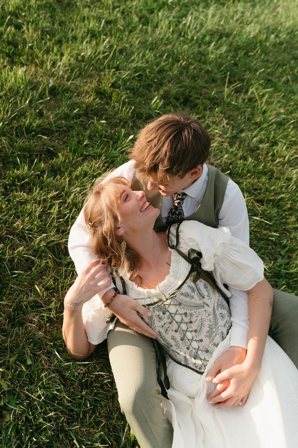 A couple in vintage-inspired clothing lies together on green grass in a romantic moment.