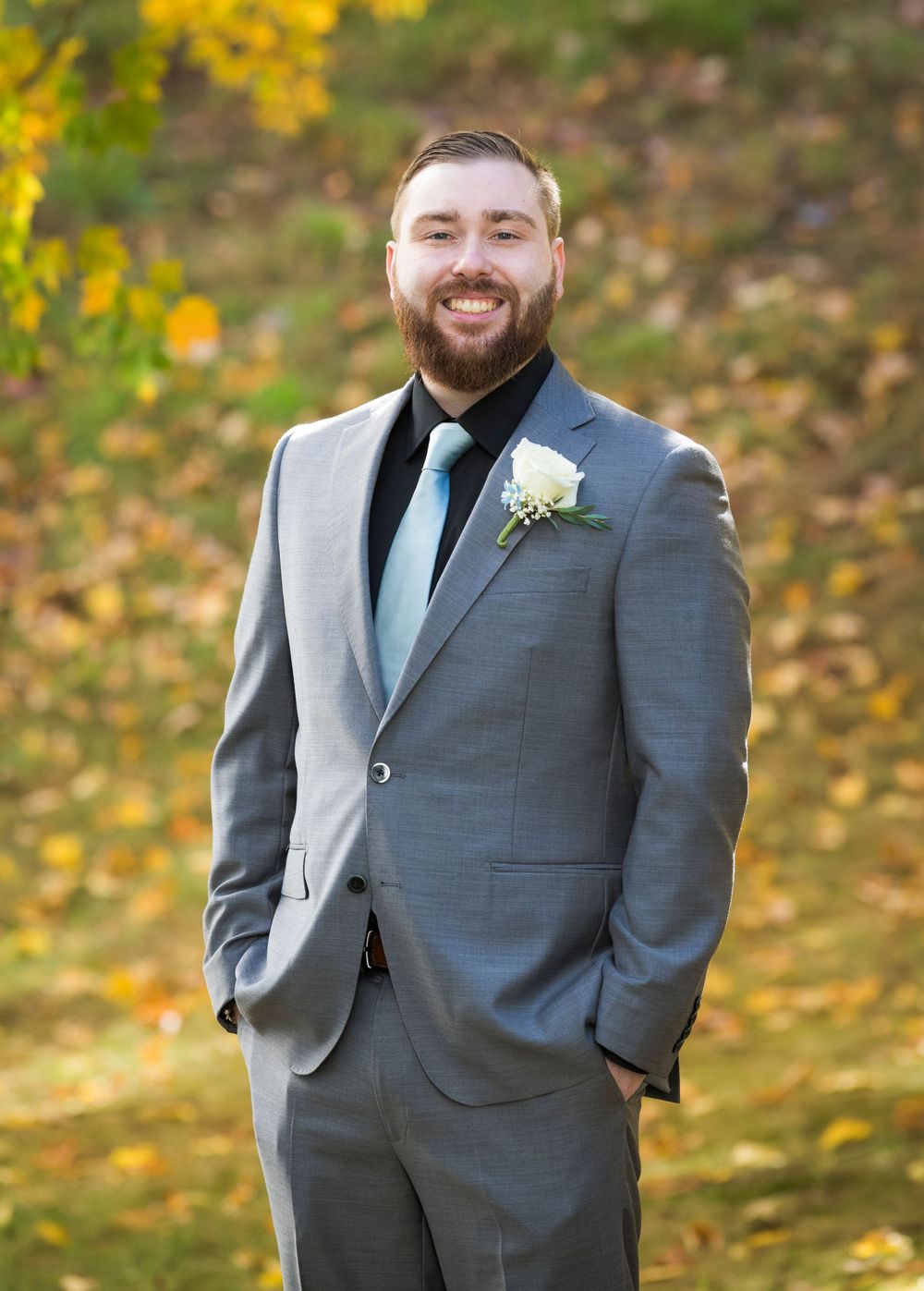 Groom in a gray suit poses outdoors against autumn foliage with yellow flowers in the background.